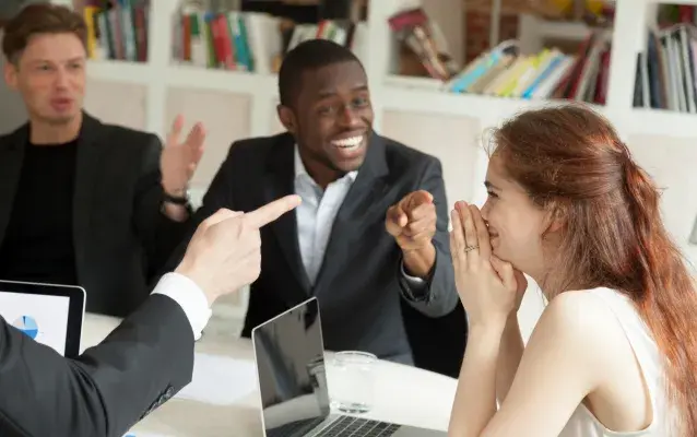 Une femme assise sur une table dans un bureau ce fait point du doigt par des hommes qui on l'air heureux. Elle fait un sourire de joie qu'elle cache timidement avec ses mains.