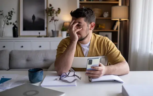 Un homme travaillant de la maison est assis à sa table avec son ordinateur et son téléphone dans les mains. Il semble découragé.