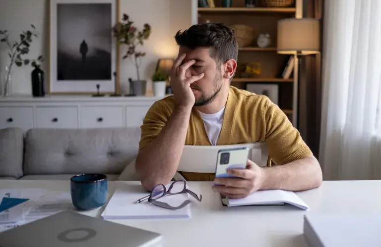 Un homme travaillant de la maison est assis à sa table avec son ordinateur et son téléphone dans les mains. Il semble découragé.