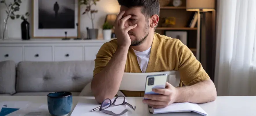 Un homme travaillant de la maison est assis à sa table avec son ordinateur et son téléphone dans les mains. Il semble découragé.